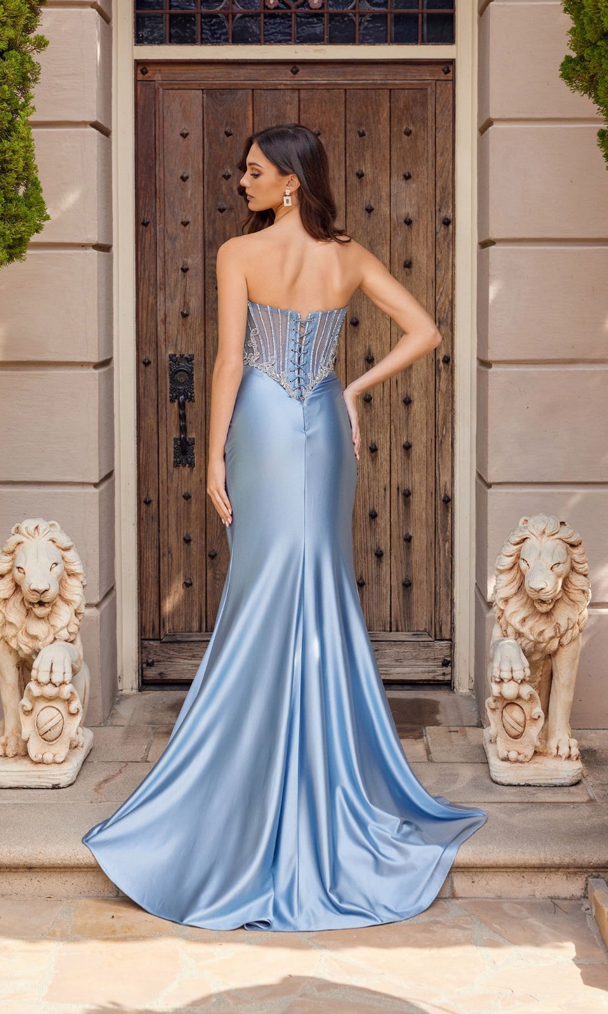 A woman in the Juliet JT2654K long prom dress with a strapless satin sheath skirt stands with her back to the camera in front of a wooden door between two stone lion statues.